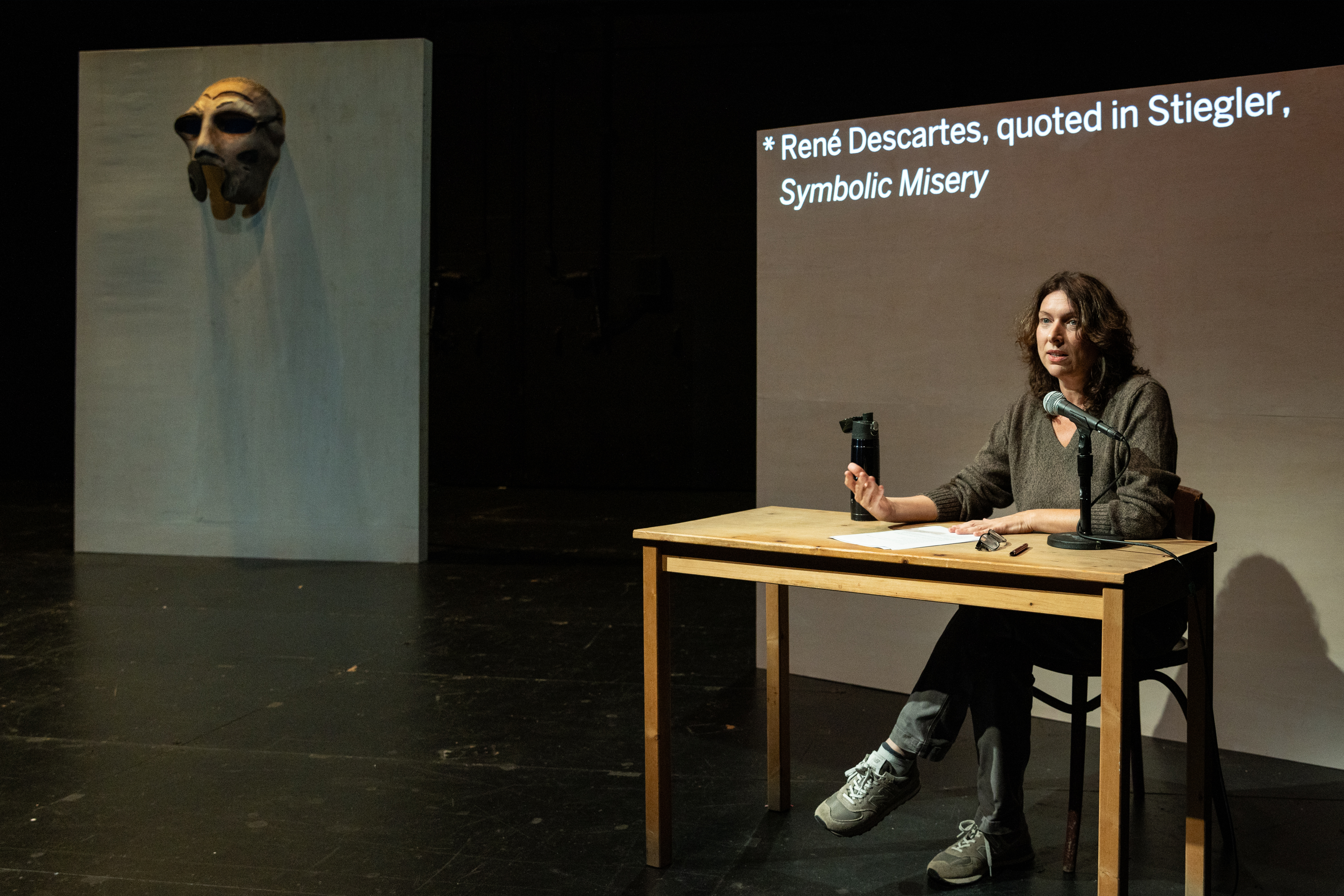 Annie Dorsen sits at a table in front of a projection screen.