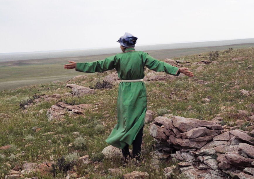 A person wearing a long sleeved green dress faces the horizon with their arms outstretched, surrounded by rocks and grass.