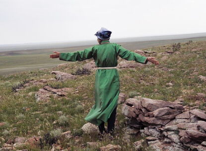 A person wearing a long sleeved green dress faces the horizon with their arms outstretched, surrounded by rocks and grass.